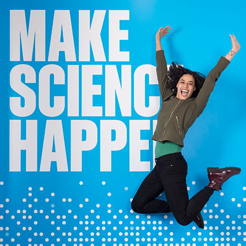A woman jumping in front of a mural that says "make science happen"